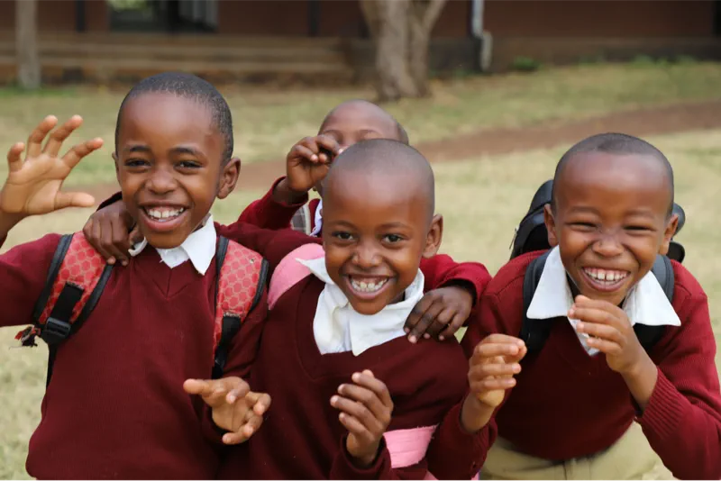 Four young school children in matching uniforms laughing and posing with their backpacks on