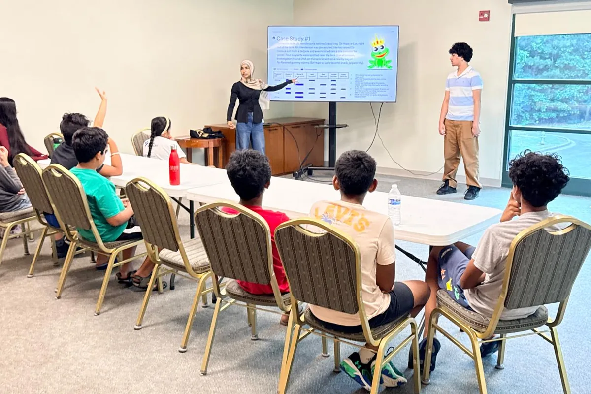 A student volunteer leading a case-study workshop for a room of younger students, one of whom has a hand raised to answer